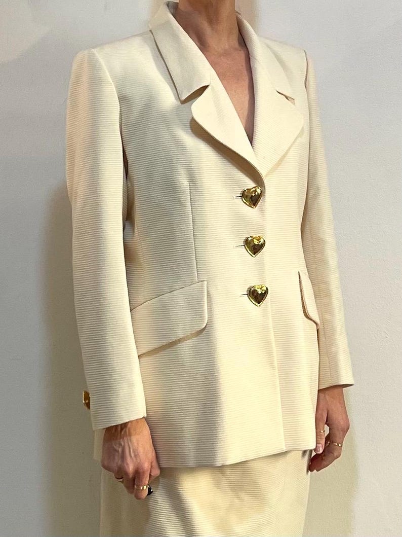 Puede incluir: Traje de blazer y falda color crema. La blazer presenta solapas con muescas, bolsillos con solapa y botones dorados en forma de coraz&oacute;n. La tela tiene una sutil textura acanalada. El traje es de estilo cl&aacute;sico y elegante.