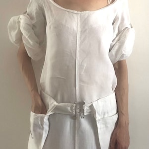 May include: A white linen dress with a scoop neck and puff sleeves. The dress has a cinched waist with a fabric belt and two large pockets on the front.