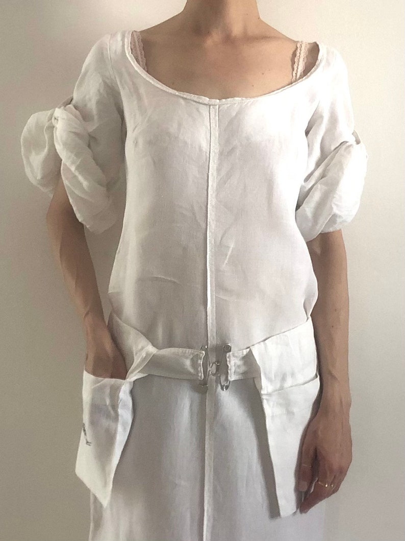 May include: A white linen dress with a scoop neck and puff sleeves. The dress has a cinched waist with a fabric belt and two large pockets on the front.