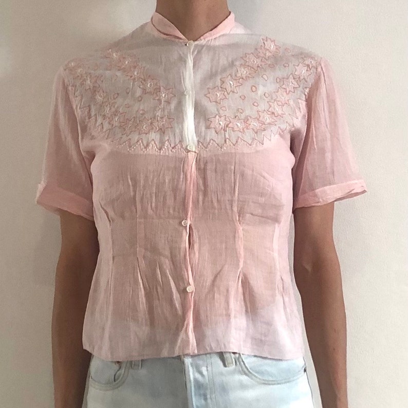 Pu&ograve; includere: Una camicia rosa pallido a maniche corte con bottoni, colletto bianco e delicati ricami rosa intorno al collo.
