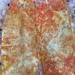 May include: Orange, yellow, and white tie-dye jeans with a floral pattern. The jeans have a small label on the back waistband that says "SPELLBOUND" in white text.