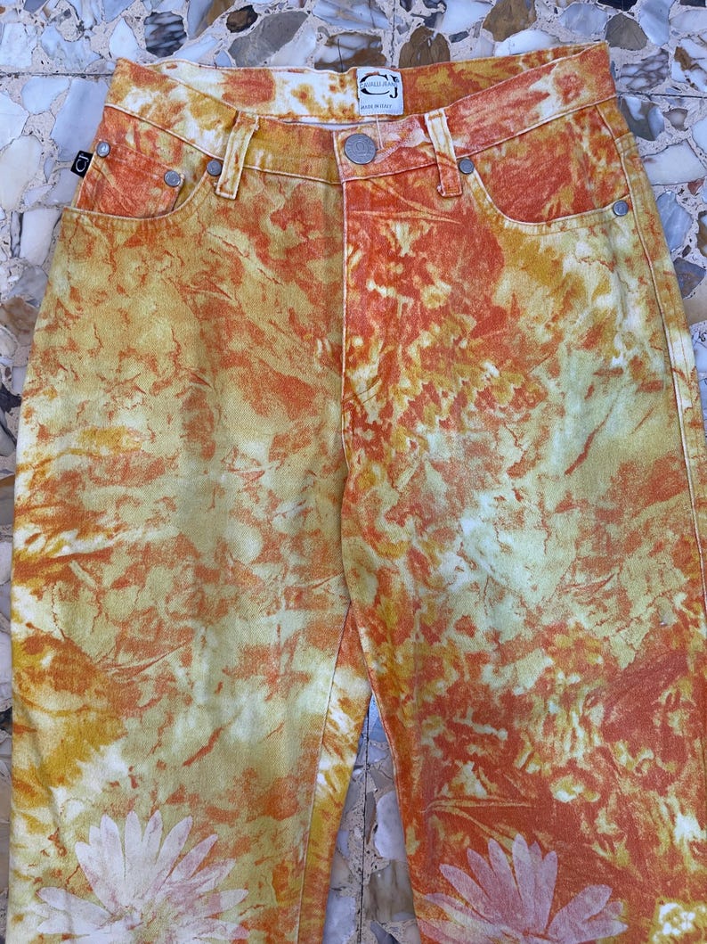 May include: Orange, yellow, and white tie-dye jeans with a floral pattern. The jeans have a small label on the back waistband that says "SPELLBOUND" in white text.