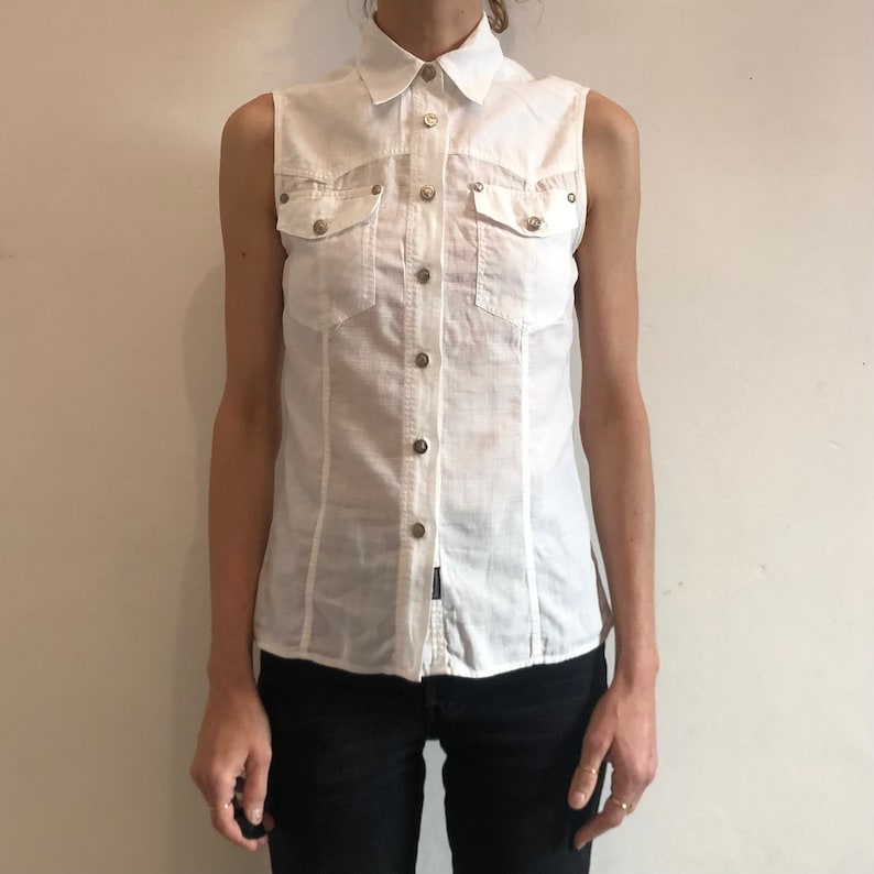 May include: A white sleeveless button-down shirt with a collared neckline and two flap pockets. The shirt has a slightly fitted silhouette.
