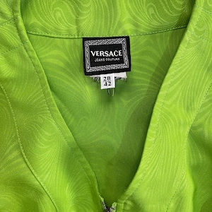 Może przedstawiać: Zielona kurtka zapinana na zamek błyskawiczny z wzorem wirującym. Kurtka ma metkę z napisem "VERSACE JEANS COUTURE" i "28 42".