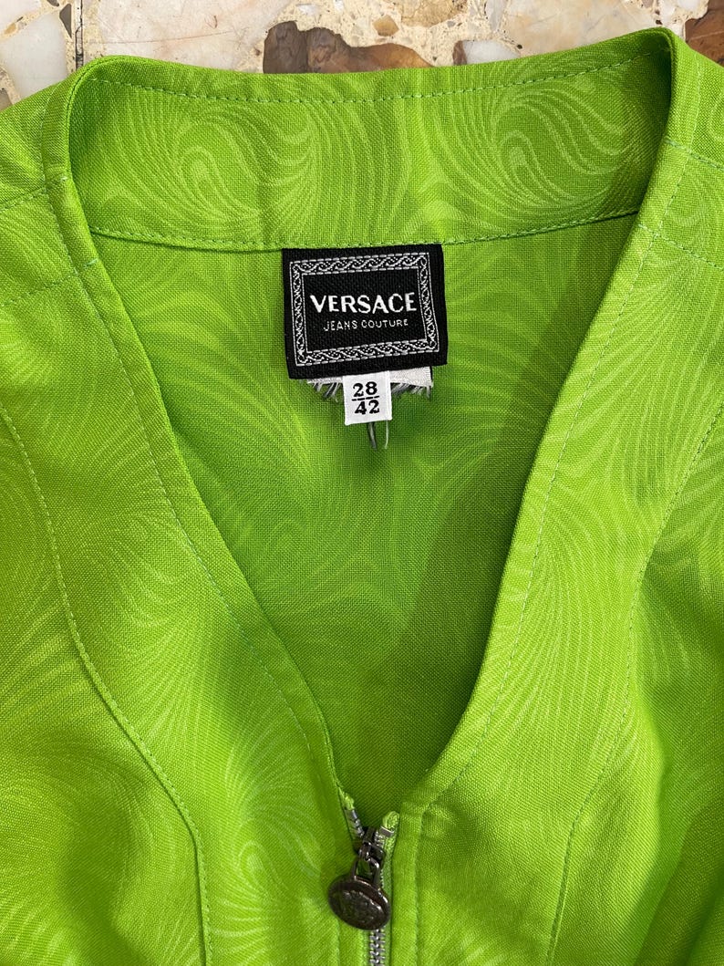 Może przedstawiać: Zielona kurtka zapinana na zamek błyskawiczny z wzorem wirującym. Kurtka ma metkę z napisem "VERSACE JEANS COUTURE" i "28 42".