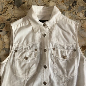 May include: A white sleeveless button-down shirt with a collared neckline and two chest pockets. The shirt has silver-toned buttons and a subtle textured fabric.
