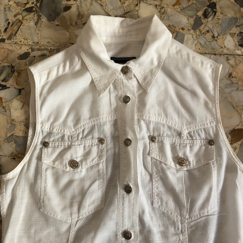 May include: A white sleeveless button-down shirt with a collared neckline and two chest pockets. The shirt has silver-toned buttons and a subtle textured fabric.