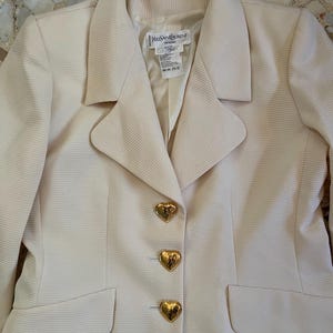 Puede incluir: Blazer color crema con botones dorados en forma de coraz&oacute;n. El blazer tiene una tela texturizada y un cuello cl&aacute;sico. La etiqueta dice "Yves Saint Laurent". Se ven dos bolsillos delanteros.