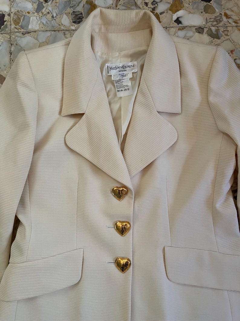 Puede incluir: Blazer color crema con botones dorados en forma de coraz&oacute;n. El blazer tiene una tela texturizada y un cuello cl&aacute;sico. La etiqueta dice "Yves Saint Laurent". Se ven dos bolsillos delanteros.