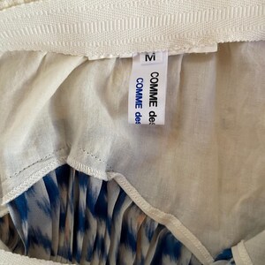 Op de afbeelding: Close-up van een rok met een witte tailleband en een blauw-wit patroon. Het label leest "M" en "COMME des GAR&Ccedil;ONS". De rok heeft een geplooid ontwerp.