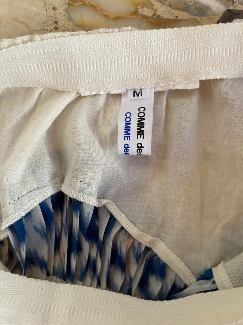 Op de afbeelding: Close-up van een rok met een witte tailleband en een blauw-wit patroon. Het label leest "M" en "COMME des GAR&Ccedil;ONS". De rok heeft een geplooid ontwerp.