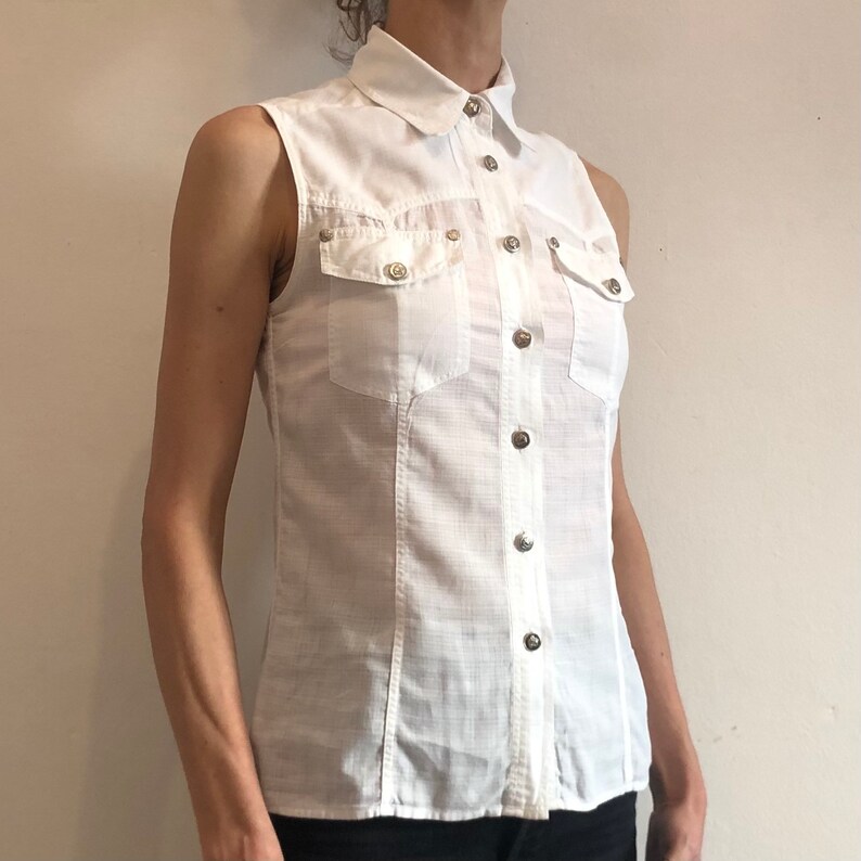 May include: A white sleeveless button-down shirt with a collared neckline and two flap pockets. The shirt has a textured fabric and silver buttons.