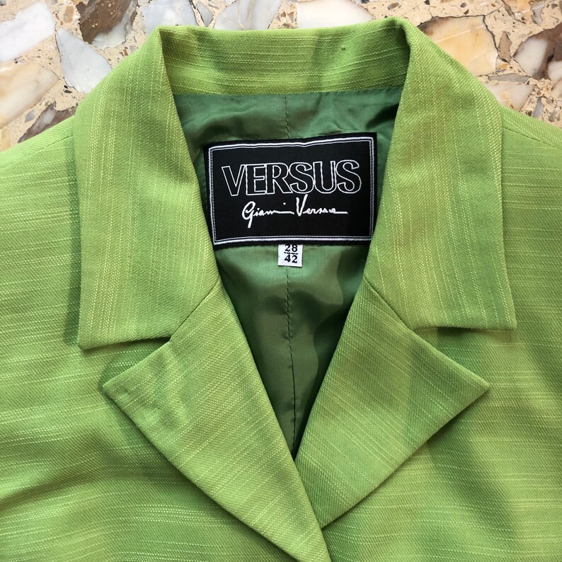 Pode incluir: Um blazer verde com uma etiqueta preta que diz "VERSUS Gianni Versace" e etiquetas de tamanho que dizem "42" e "28".