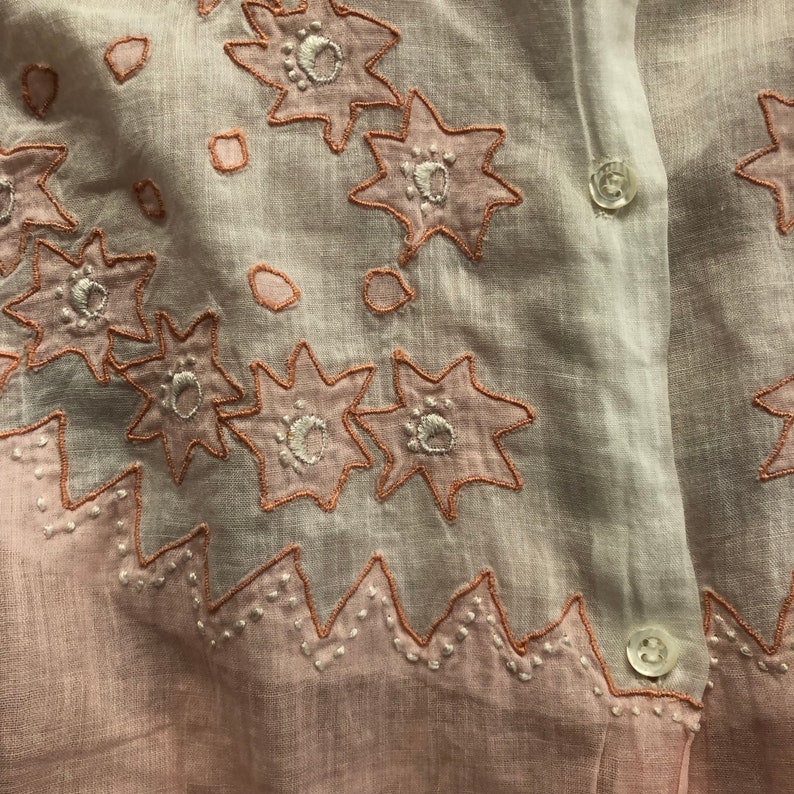 Pu&ograve; includere: Una camicia di cotone rosa e bianca con fiori ricamati. I fiori sono bordati di rosa e hanno centri bianchi. La camicia ha una chiusura con bottoni sul davanti e un orlo a festoni.