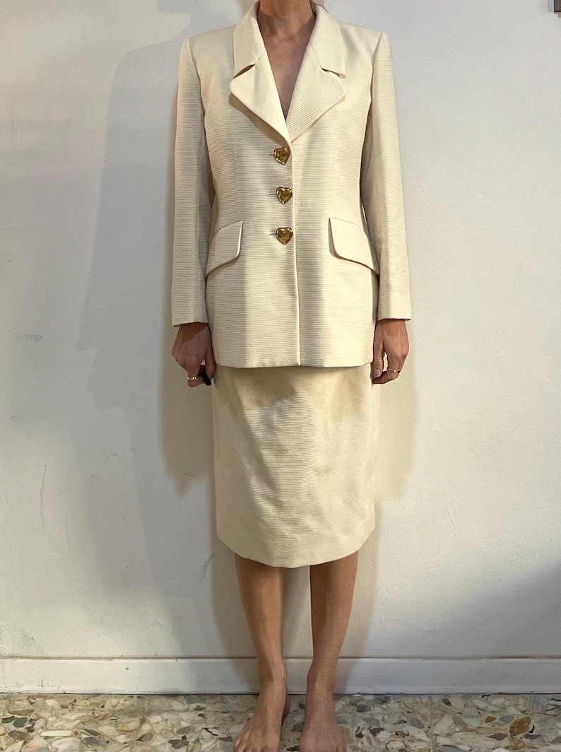 Puede incluir: Traje de falda color crema. La chaqueta tiene solapas, dos bolsillos delanteros y tres botones dorados en forma de coraz&oacute;n. La falda llega a la rodilla. El traje est&aacute; hecho de una tela texturizada.