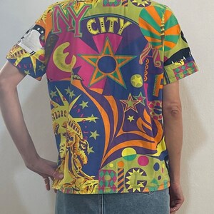 K&ouml;nnte beinhalten: Ein kurz&auml;rmeliges Hemd mit Kn&ouml;pfen und einem bunten, psychedelischen Druck mit den Worten "NY CITY" und Bildern der Freiheitsstatue, Sternen und anderen Designs zum Thema New York City.