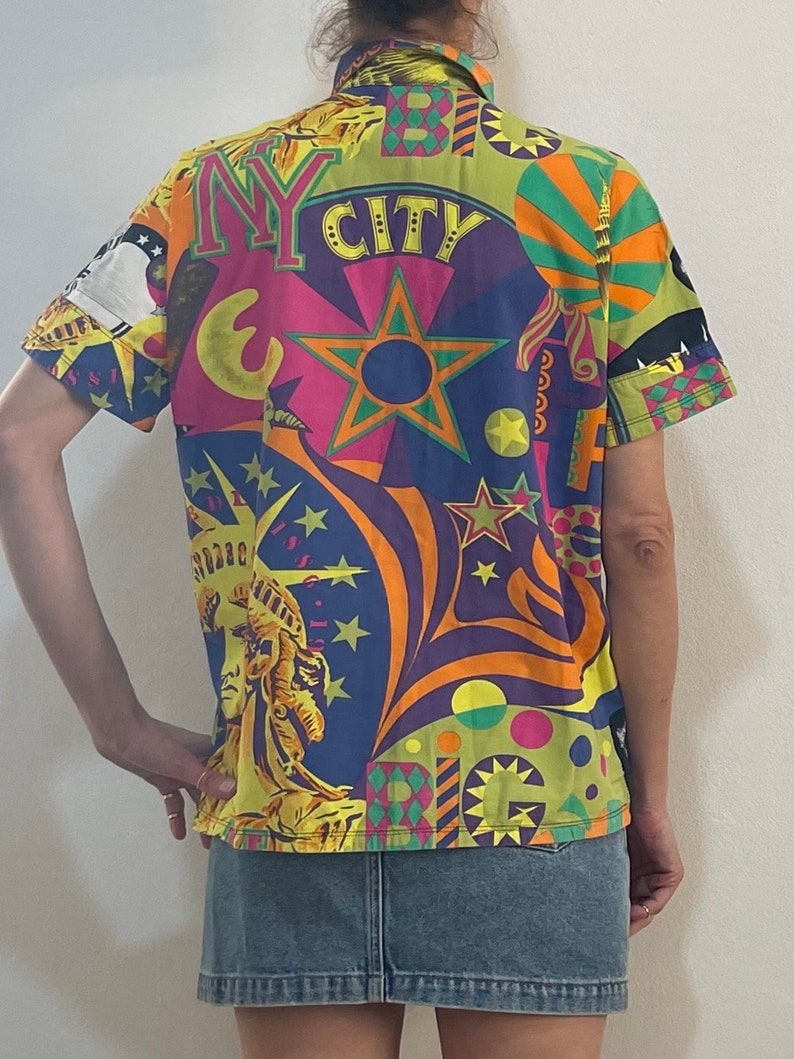 K&ouml;nnte beinhalten: Ein kurz&auml;rmeliges Hemd mit Kn&ouml;pfen und einem bunten, psychedelischen Druck mit den Worten "NY CITY" und Bildern der Freiheitsstatue, Sternen und anderen Designs zum Thema New York City.