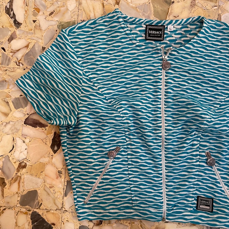 Op de afbeelding: Een blauw en wit gestreept getailleerd jack met een zilveren rits en zilveren hardware. Het jack heeft het woord "VERSACE" op het label.