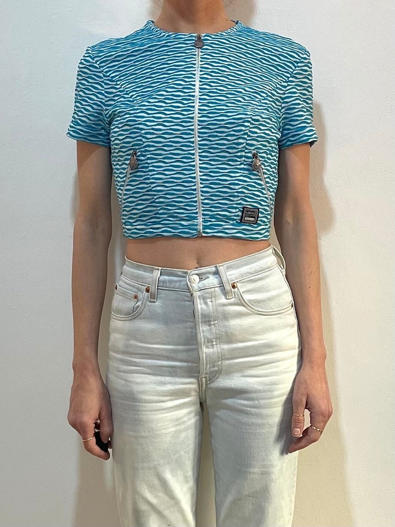 Op de afbeelding: Een blauw en wit gestreept getailleerd jack met een ritssluiting en een klein zakje aan de voorkant. Het jack heeft een Versace-logo op het zakje.