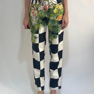 May include: High-waisted pants featuring a bold black and white checkerboard pattern across the legs. The upper hip area is decorated with a vibrant, detailed botanical print of green palm leaves, yellow hibiscus, and white orchid flowers.