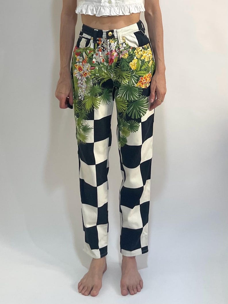May include: High-waisted pants featuring a bold black and white checkerboard pattern across the legs. The upper hip area is decorated with a vibrant, detailed botanical print of green palm leaves, yellow hibiscus, and white orchid flowers.