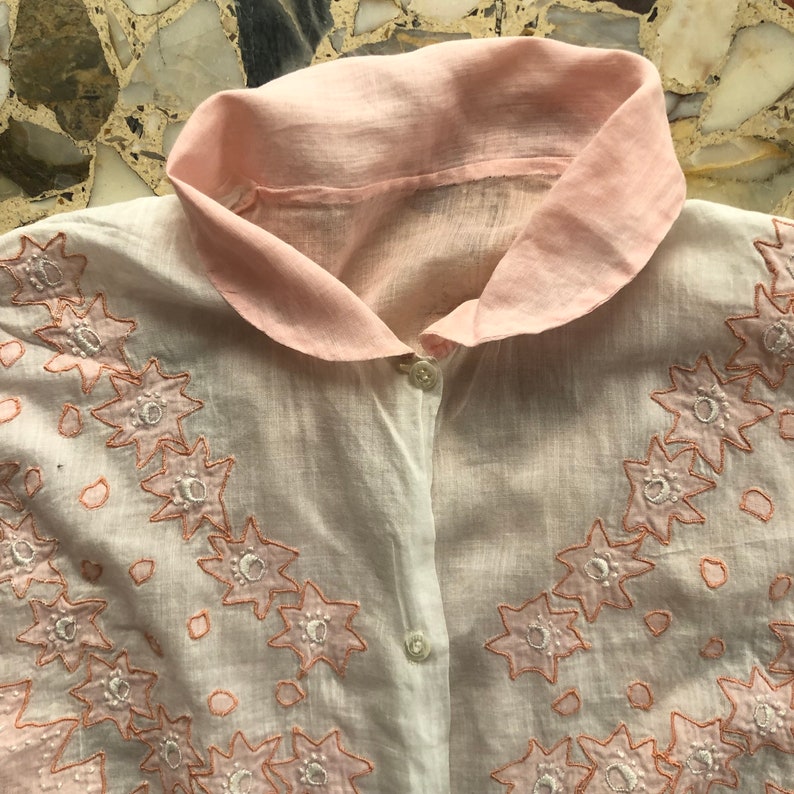 Pu&ograve; includere: Una camicia di cotone bianco con colletto rosa e ricamo floreale delicato. Il ricamo &egrave; di colore rosa chiaro e presenta un motivo ripetitivo di piccoli fiori.