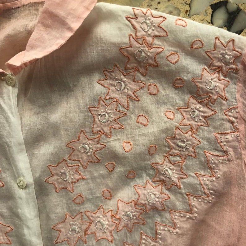 Pu&ograve; includere: Una camicia di cotone rosa e bianca con fiori bianchi ricamati con contorno rosa.
