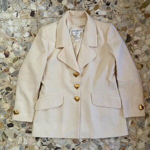 Puede incluir: Una blazer color crema con tejido texturizado. El blazer presenta botones dorados en forma de coraz&oacute;n y detalles a juego en los pu&ntilde;os. La etiqueta dice "Yves Saint Laurent".