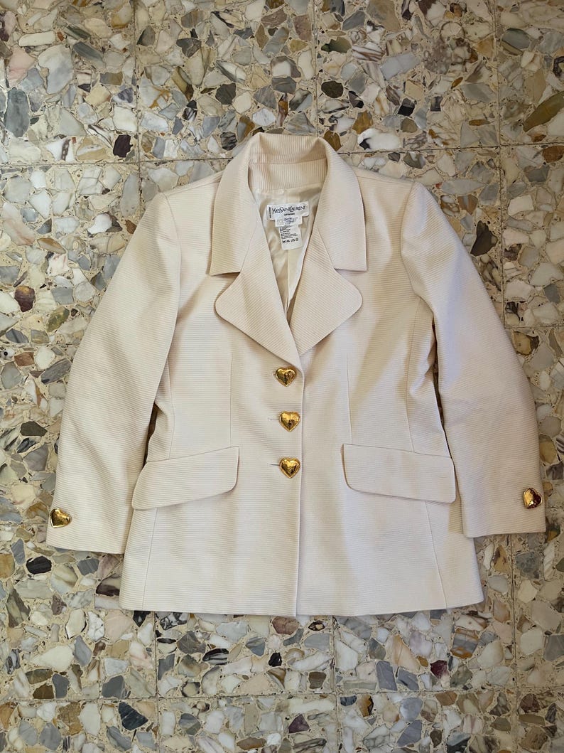 Puede incluir: Una blazer color crema con tejido texturizado. El blazer presenta botones dorados en forma de coraz&oacute;n y detalles a juego en los pu&ntilde;os. La etiqueta dice "Yves Saint Laurent".