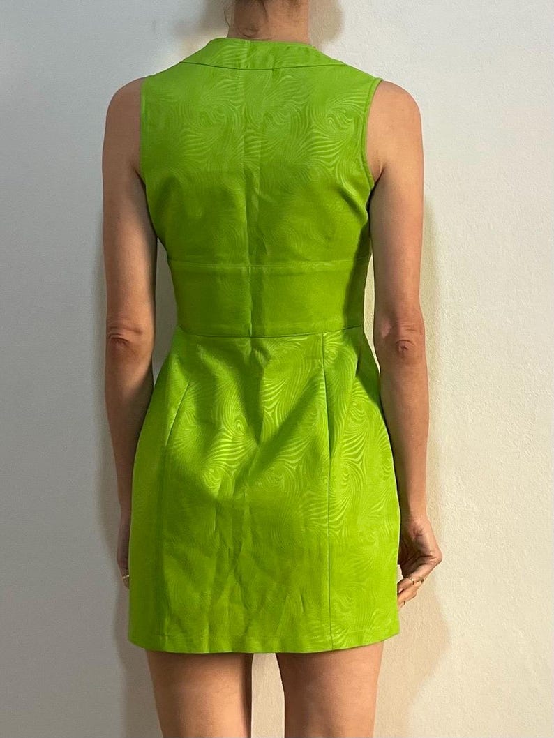 Może przedstawiać: Zielona sukienka bez rękaw&oacute;w z wirującym wzorem. Sukienka ma dekolt w kształcie litery V i dopasowany kr&oacute;j.