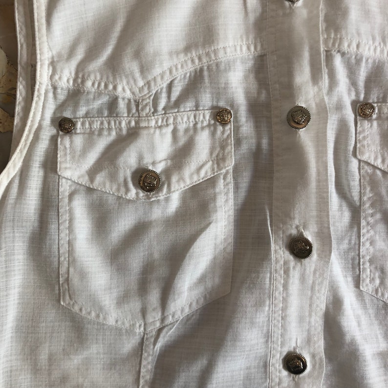 May include: White sleeveless shirt with a button-up front and a chest pocket. The shirt has silver buttons with a raised design.