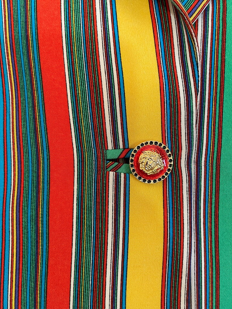 May include: A close-up of a striped fabric with a gold button featuring a lion's head design. The fabric is a mix of red, yellow, green, blue, and white stripes.