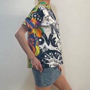 K&ouml;nnte beinhalten: Ein wei&szlig;es, kurz&auml;rmeliges Hemd mit Kn&ouml;pfen und einem bunten, grafischen Druck mit den Worten "Love" und "New York" sowie Bildern von Sternen und der Freiheitsstatue. Das Hemd wird mit einem Jeansrock getragen.