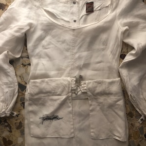 May include: White linen dress with long sleeves and a scoop neck. The dress has a small pocket on the front with the text "Gasoline Jeans" embroidered in black. The dress has a brown label with the text "Gasoline Jeans" on the front.
