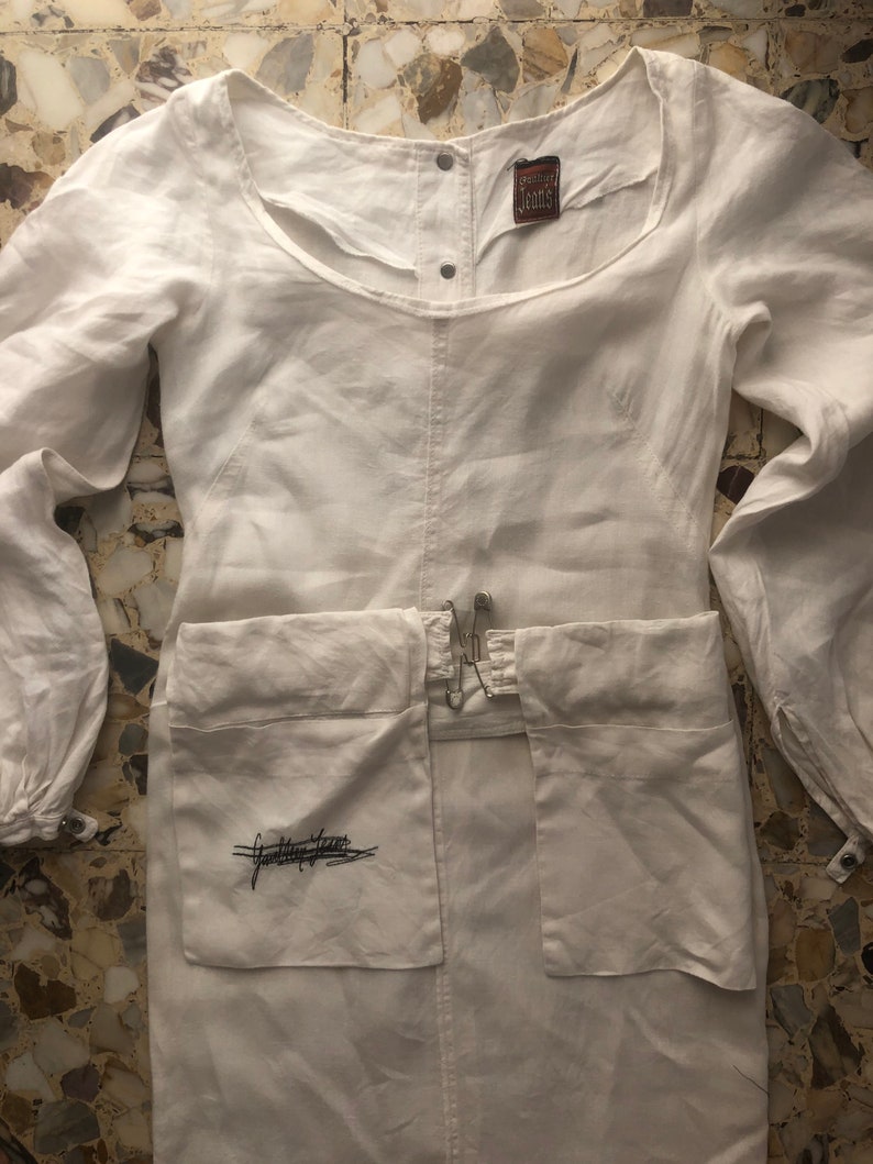 May include: White linen dress with long sleeves and a scoop neck. The dress has a small pocket on the front with the text "Gasoline Jeans" embroidered in black. The dress has a brown label with the text "Gasoline Jeans" on the front.