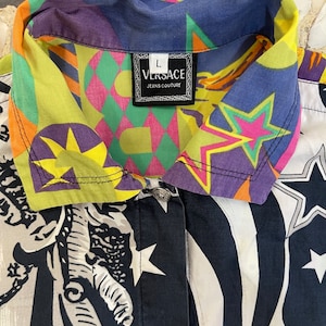 K&ouml;nnte beinhalten: Ein mehrfarbiges Hemd mit Kn&ouml;pfen, geometrischem Muster und Sternen. Das Hemd hat einen schwarz-wei&szlig;en Druck einer Figur auf der Vorderseite. Das Etikett auf dem Hemd lautet "VERSACE JEANS COUTURE".
