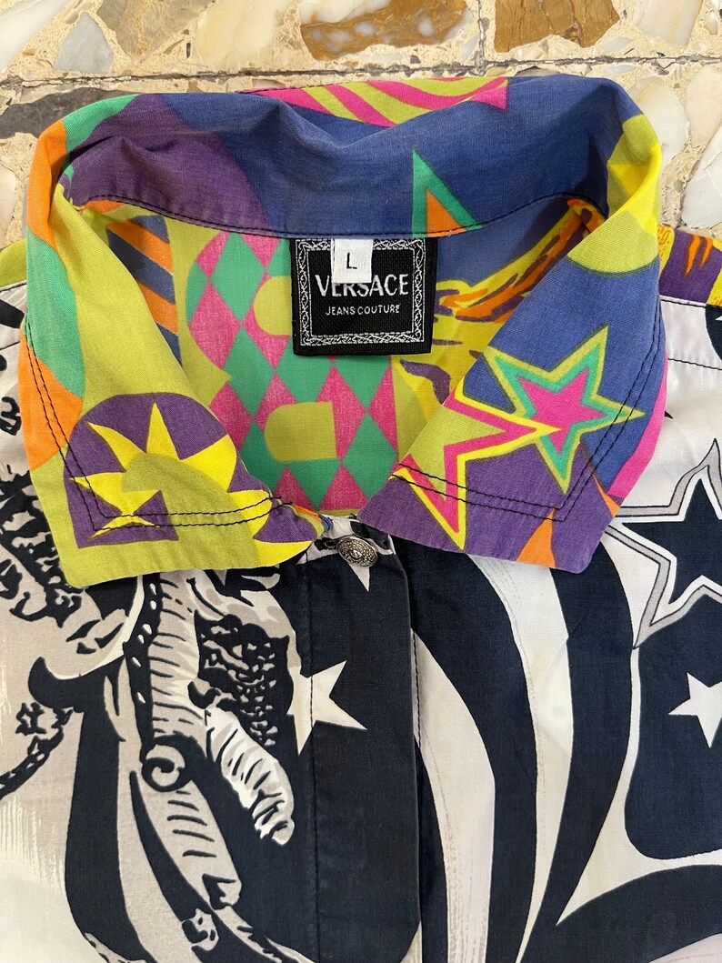 K&ouml;nnte beinhalten: Ein mehrfarbiges Hemd mit Kn&ouml;pfen, geometrischem Muster und Sternen. Das Hemd hat einen schwarz-wei&szlig;en Druck einer Figur auf der Vorderseite. Das Etikett auf dem Hemd lautet "VERSACE JEANS COUTURE".