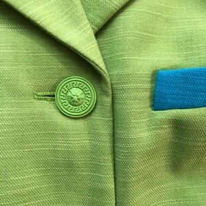 Pode incluir: Um blazer verde com um bot&atilde;o verde com um design de cabe&ccedil;a de le&atilde;o. O blazer tem um bolso azul no lado direito.