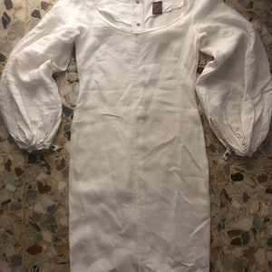 May include: A white linen dress with long sleeves and a gathered neckline. The dress has a button closure at the neck and a small label with the text "Vintage Linen" on the front.