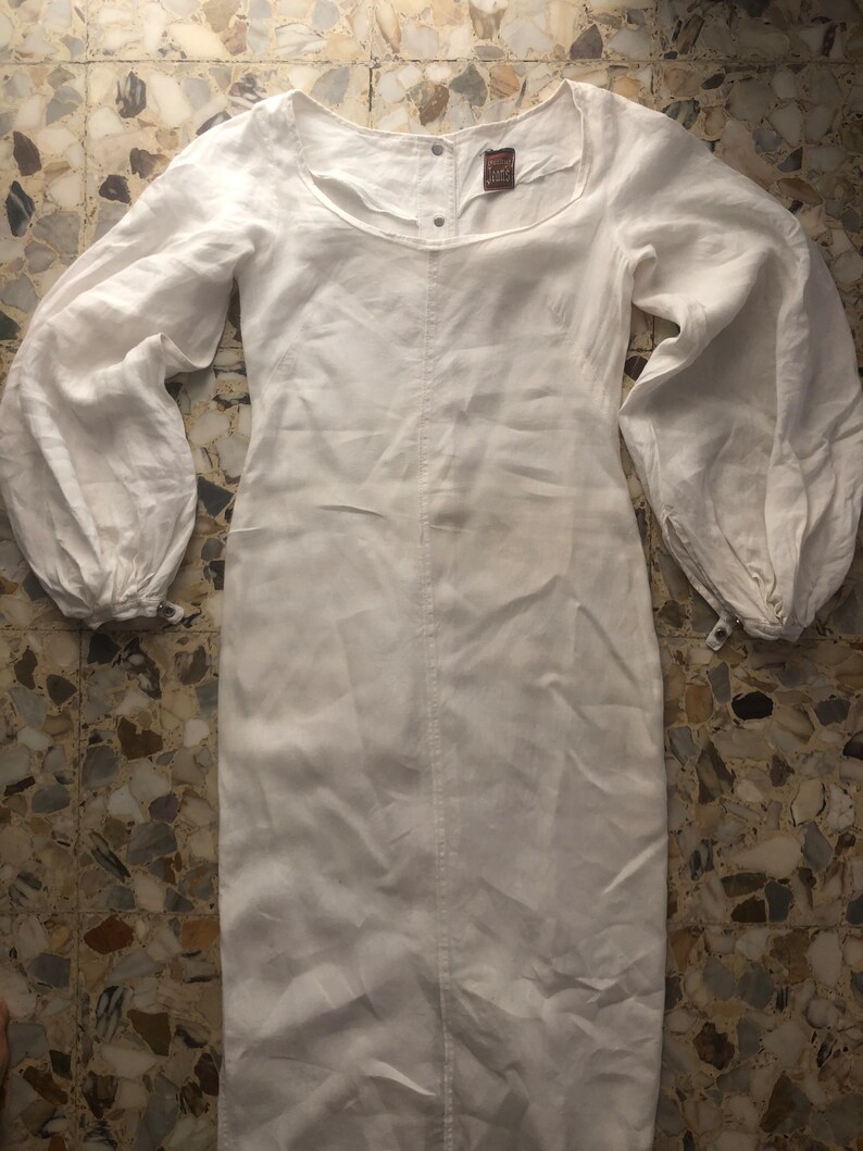 May include: A white linen dress with long sleeves and a gathered neckline. The dress has a button closure at the neck and a small label with the text "Vintage Linen" on the front.