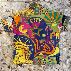 K&ouml;nnte beinhalten: Ein kurz&auml;rmeliges Hemd mit Kn&ouml;pfen und einem farbenfrohen, psychedelischen Druck mit den Worten "NY City" und "Big Apple". Der Druck zeigt auch eine Darstellung der Freiheitsstatue und anderer ikonischer Bilder von New York City.