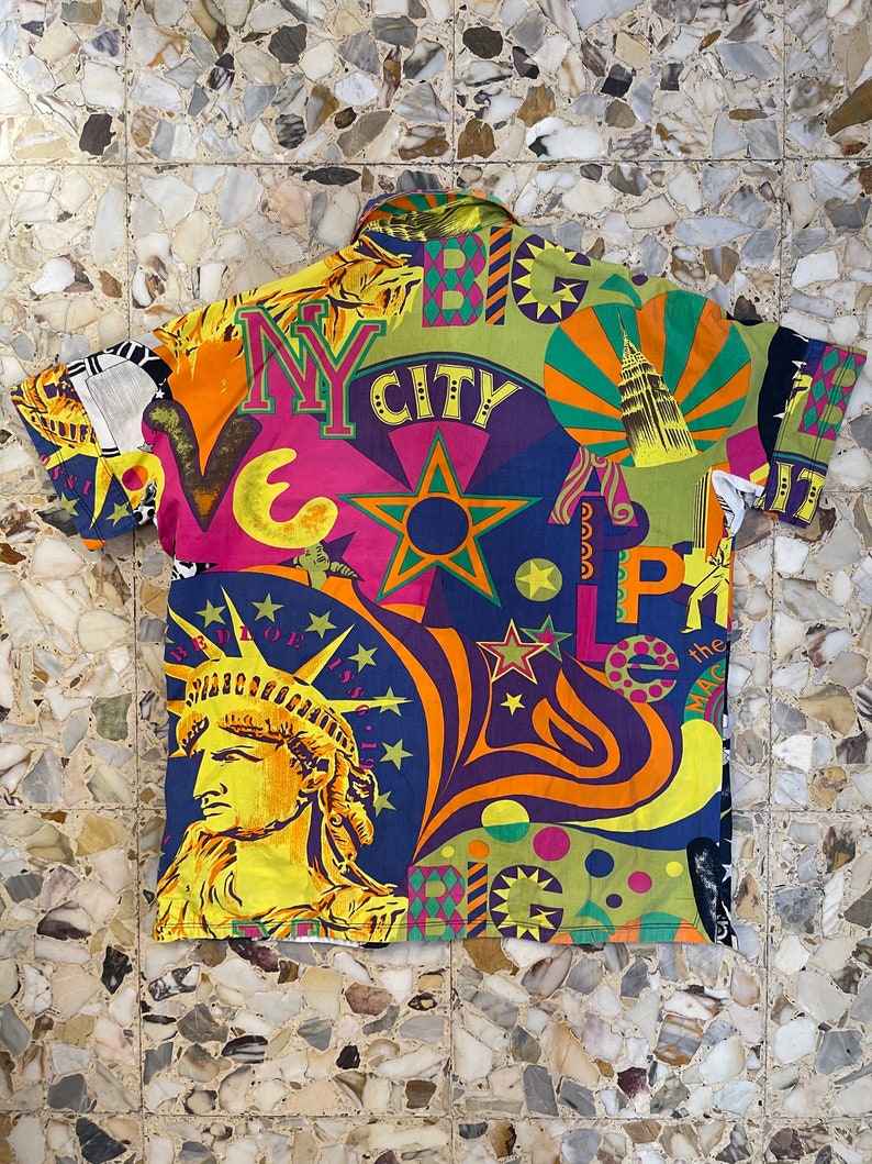 K&ouml;nnte beinhalten: Ein kurz&auml;rmeliges Hemd mit Kn&ouml;pfen und einem farbenfrohen, psychedelischen Druck mit den Worten "NY City" und "Big Apple". Der Druck zeigt auch eine Darstellung der Freiheitsstatue und anderer ikonischer Bilder von New York City.