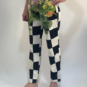 May include: High-waisted pants featuring a bold black and white checkered pattern. The upper front section is decorated with a vibrant floral print in shades of yellow, pink, and green, depicting tropical flowers and palm leaves on the fabric.