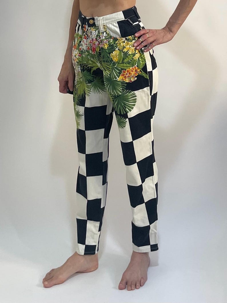 May include: High-waisted pants featuring a bold black and white checkered pattern. The upper front section is decorated with a vibrant floral print in shades of yellow, pink, and green, depicting tropical flowers and palm leaves on the fabric.