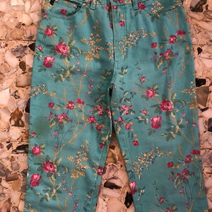 May include: A pair of blue denim jeans with a floral print featuring pink and red roses, green leaves, and yellow stems. The jeans have a high waist and a straight leg.