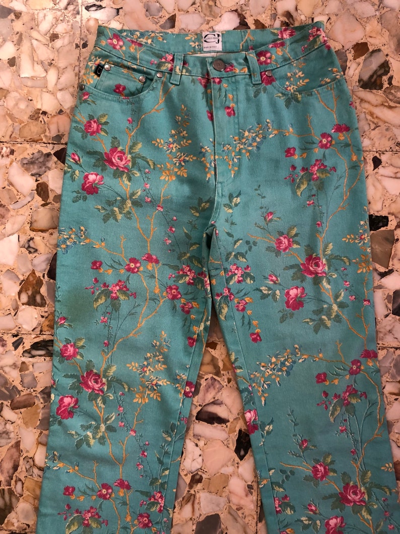 May include: A pair of blue denim jeans with a floral print featuring pink and red roses, green leaves, and yellow stems. The jeans have a high waist and a straight leg.
