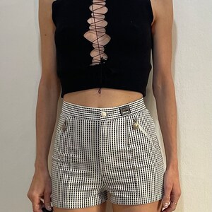 May include: Black sleeveless top with a lace-up front paired with high-waisted shorts. The shorts feature a black and white checkered pattern, a button closure, and a small zipper pocket. The outfit is a stylish ensemble.