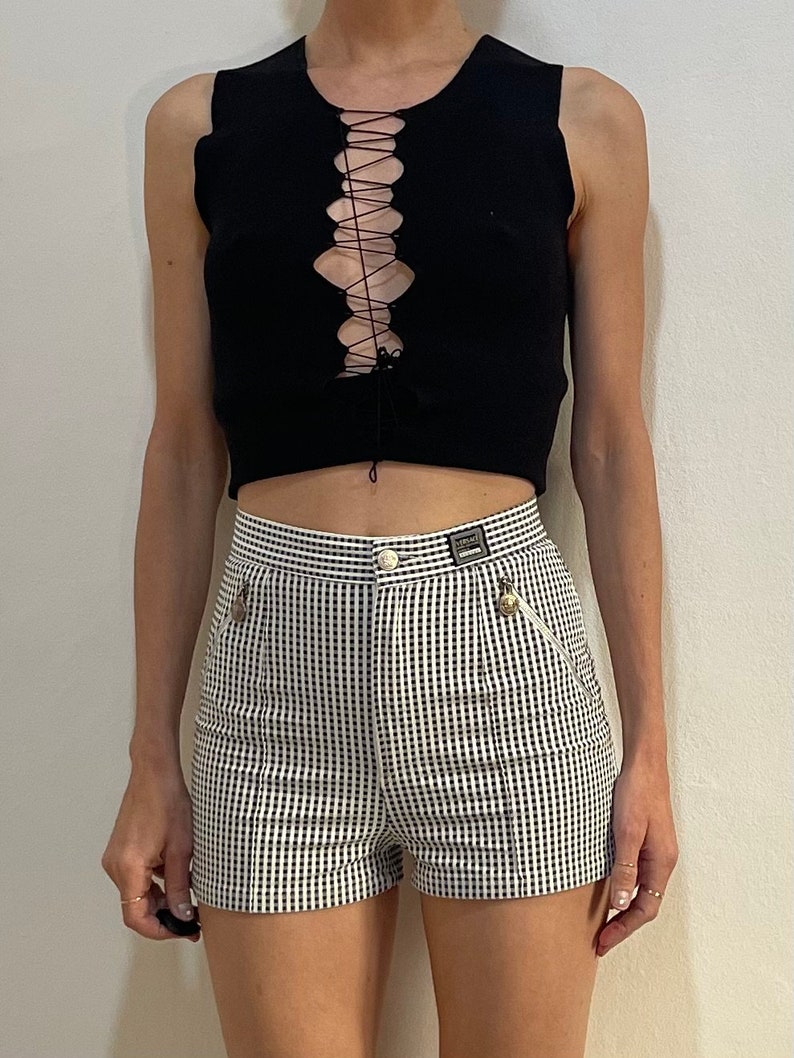 May include: Black sleeveless top with a lace-up front paired with high-waisted shorts. The shorts feature a black and white checkered pattern, a button closure, and a small zipper pocket. The outfit is a stylish ensemble.