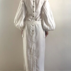 May include: A white linen dress with long sleeves and a gathered skirt. The dress has a button-up back and a belt.