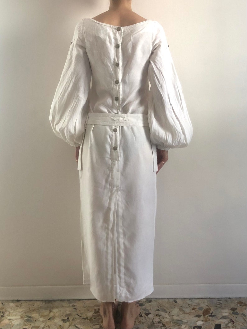 May include: A white linen dress with long sleeves and a gathered skirt. The dress has a button-up back and a belt.